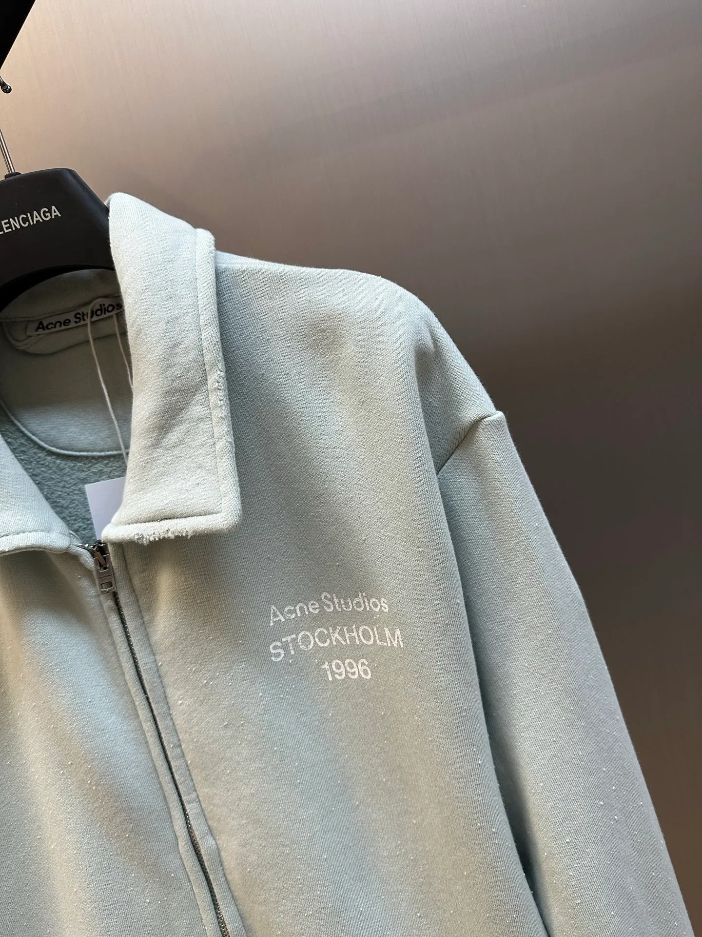 Ac vintage collared zipper sweatshirt uses 32-count top layer and 40-count bottom layer of 3-strand composite fleece structure, with a weight of g/㎡. The 32-count yarn on the top layer is fine and smooth, giving the surface a smooth touch and excellent anti-pilling properties; the 40-count 3-ply yarn on the bottom layer is treated with a raising process to form a rich and soft fluff layer, providing excellent warmth and skin-friendly wrapping. With 21 2×2 ribs, the g/㎡ weight ensures that the collar, cuffs and hem have excellent tensile fatigue resistance and shape retention capabilities. During piece dyeing, the entire fabric is immersed in the dye vat after weaving, and the dye molecules fully penetrate into the fibers, ensuring full, deep colors and excellent color fastness. Each batch of dyeing produces subtle differences, making the color tone of the single product unique. On this basis, the paint is sprayed irregularly on the surface of the garment through manual spray painting to create a splash-like retro effect. The distressed processing of holes causes the color to fade and mottle in parts. The ribbed trim is also treated to form slight whitening and fluffing, which visually echoes the spray-painted effect on the body, making the new clothes appear as if they have been worn for a long time and exposed to the sun. The front logo stamp uses a slurry printing process. The printing slurry contains a special microcapsule foaming agent. After being baked at high temperature, the slurry layer expands to form three-dimensional bulges, and the surface presents a granular texture similar to inkjet paint. This process simulates the effect of random drops of pigment after being atomized during spray gun spraying. The edges of the pattern present a natural feathering transition, rather than the rigid boundaries of glue printing. The printing surface has a fine graininess and soft elasticity to the touch, forming a closed loop of the process with the distressed effect of the spray painting on the body. size：xs s m l